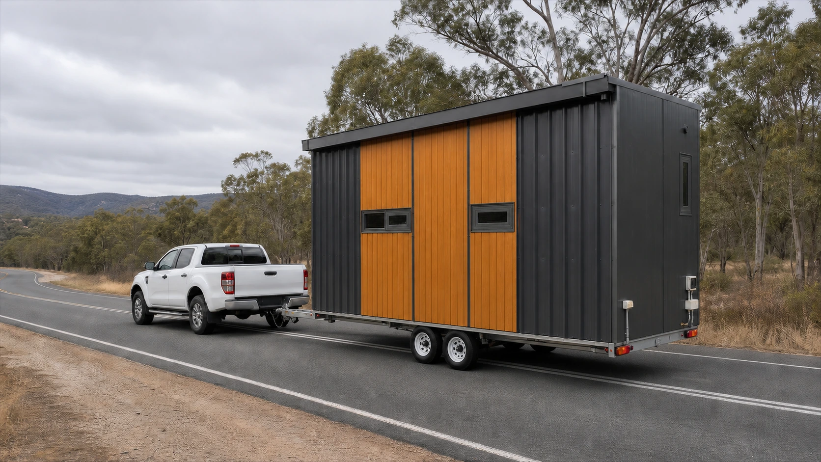 A completed tiny house with dark cladding and timber-clad end wall, on a tandem-axle trailer towed by a white dual-cab ute along a country road.