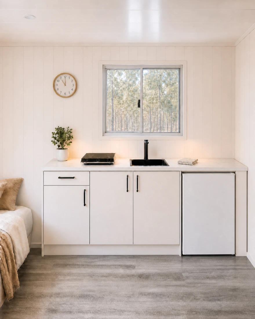 Kitchenette straight-on: white cabinetry with black handles under a stone benchtop, induction cooktop, black sink and tap below a sliding window onto bushland, an under-bench fridge to the right and the bed edge at left.