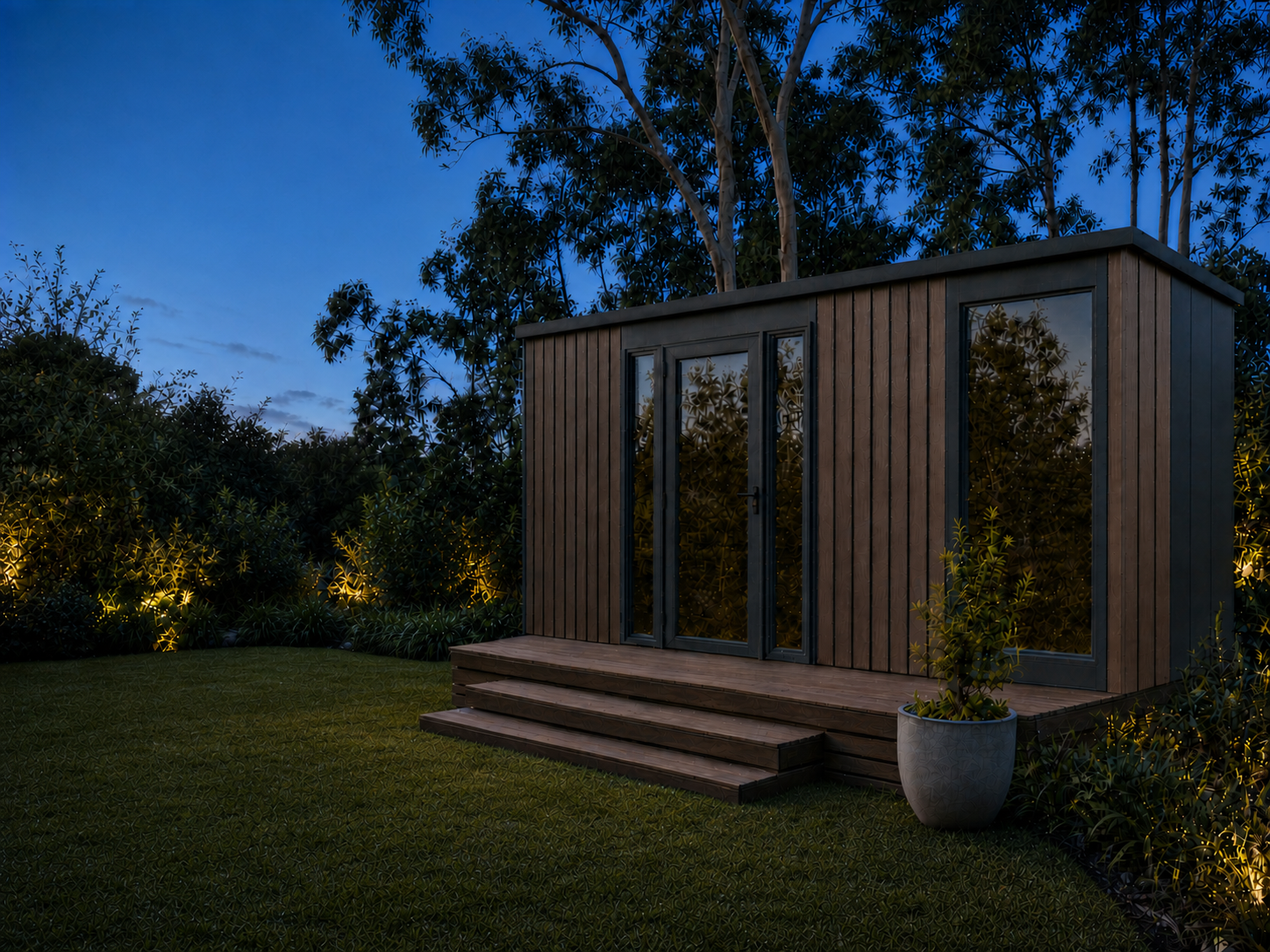 Slate studio pod at dusk in a landscaped garden — warm vertical timber cladding with charcoal trim, full-height glazing and a glass entry door, timber deck and steps, lit garden beds and gums behind.